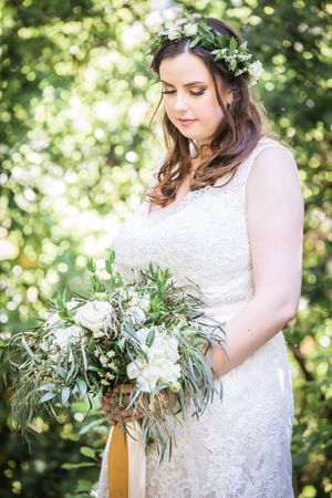 Bride in lace dress with flower crown holds elegant white and green wedding bouquet.