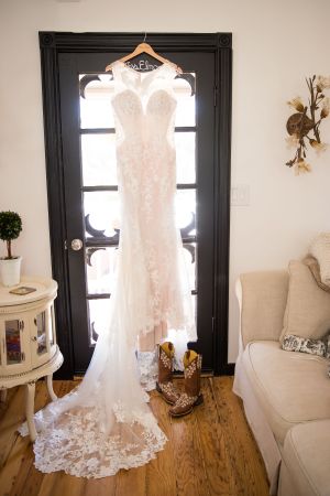 Elegant lace wedding dress hanging on display against a black door with cowboy boots below.