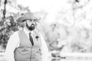 A black and white portrait of a groomsman in a tan vest and cowboy hat at an outdoor wedding.