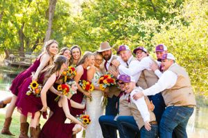 Fun wedding party group photo with burgundy dresses and sunflower bouquets by a pond.