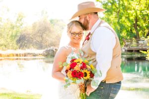 A rustic outdoor wedding couple sharing a moment by a pond with a vibrant sunflower bouquet.