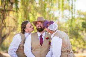 Wedding party members in tan vests sharing a candid moment under willow trees.