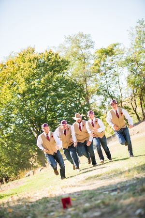 Groomsmen in matching vests running across a sunlit field during wedding festivities.