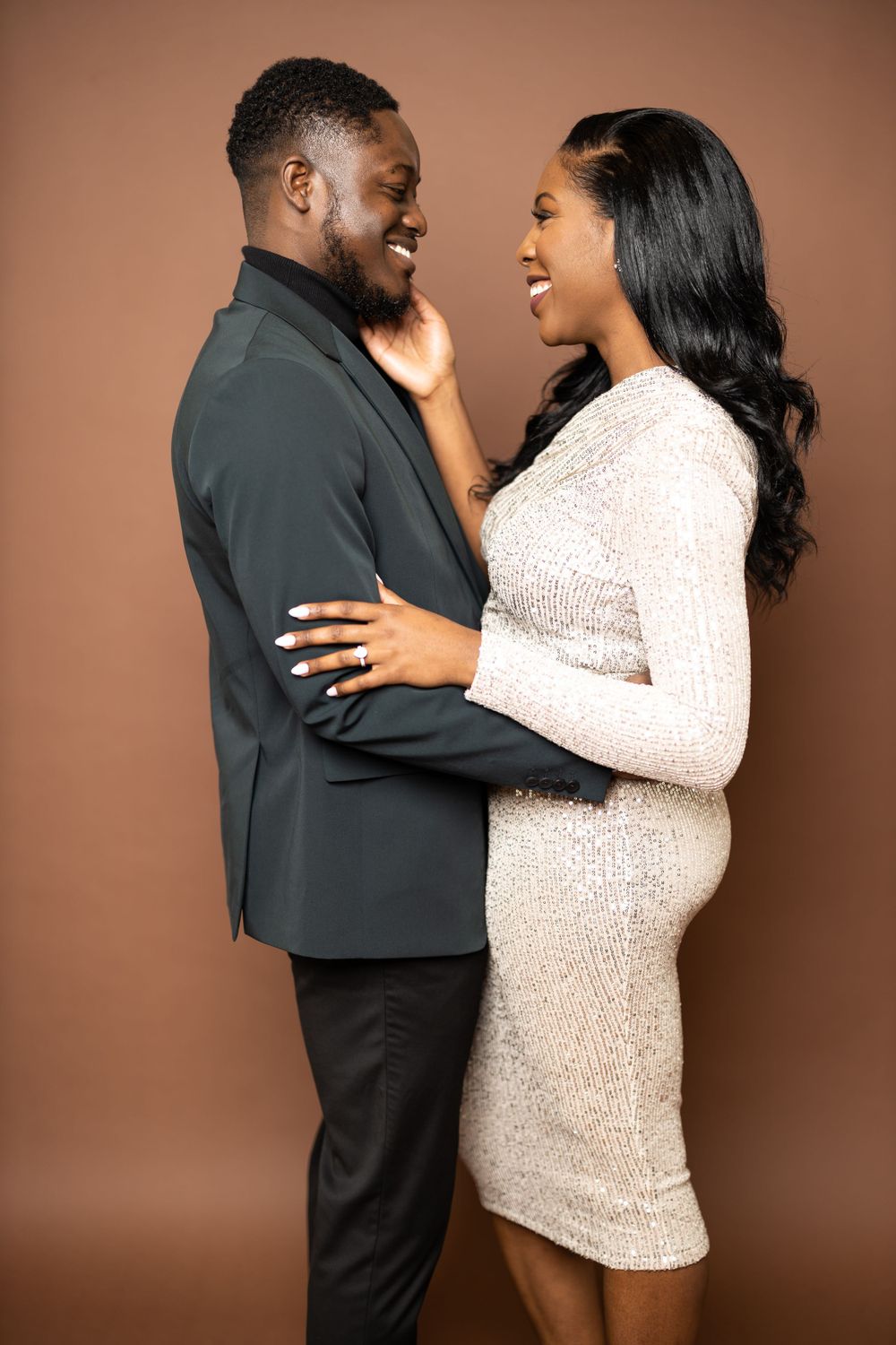 A couple shares an intimate moment in formal attire against a brown studio backdrop.