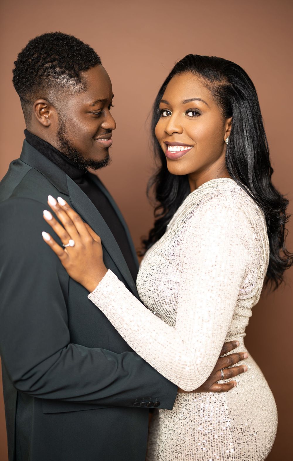 Engaged couple embraces in formal attire with sparkly white dress and black suit against brown background.