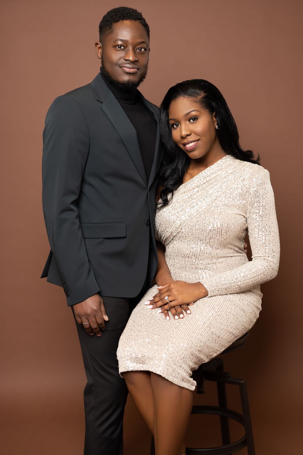 A couple poses in coordinated formal wear for an engagement photo against a brown backdrop.