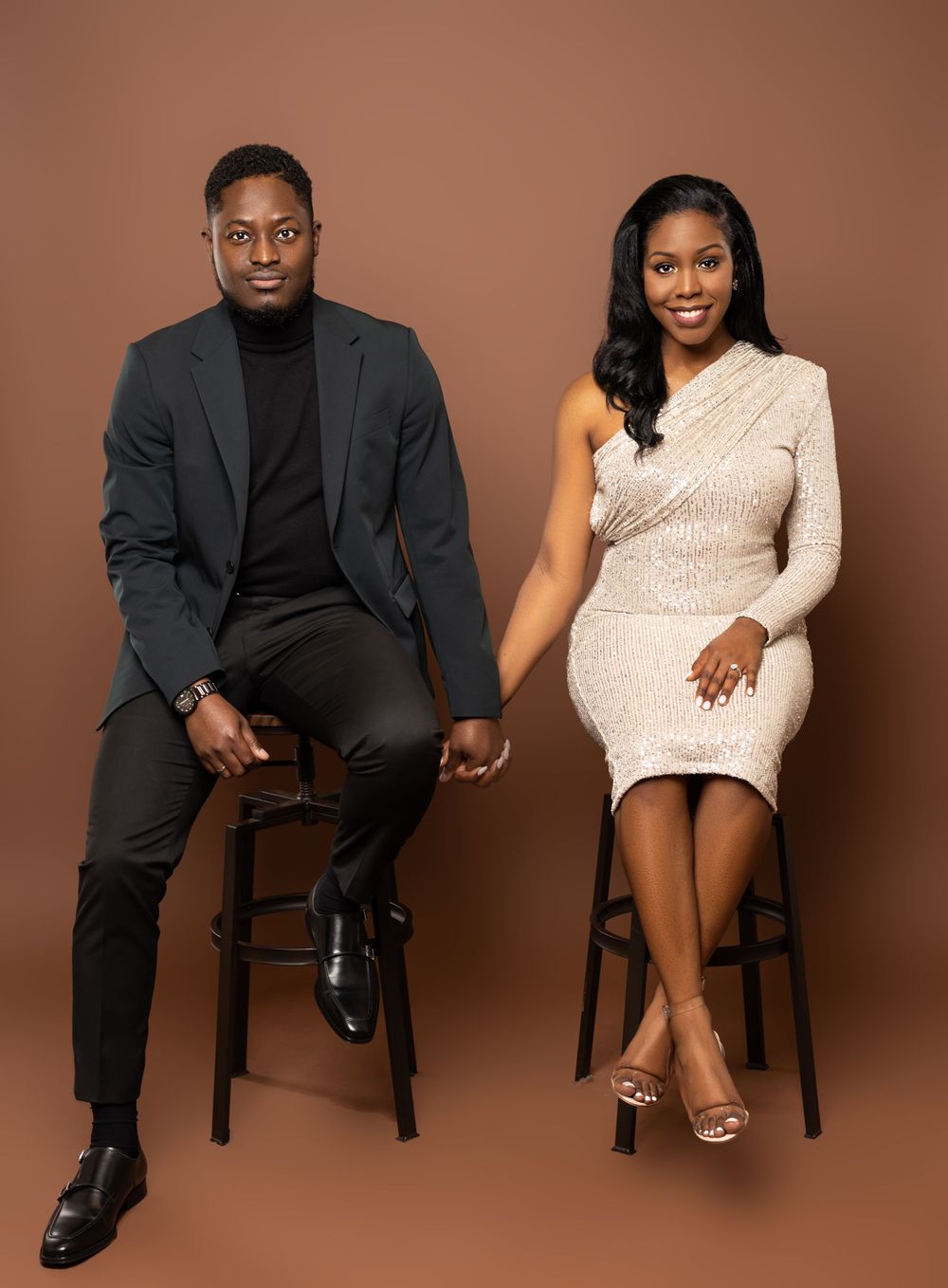 A couple sits on bar stools in coordinated formal wear against a brown studio backdrop.