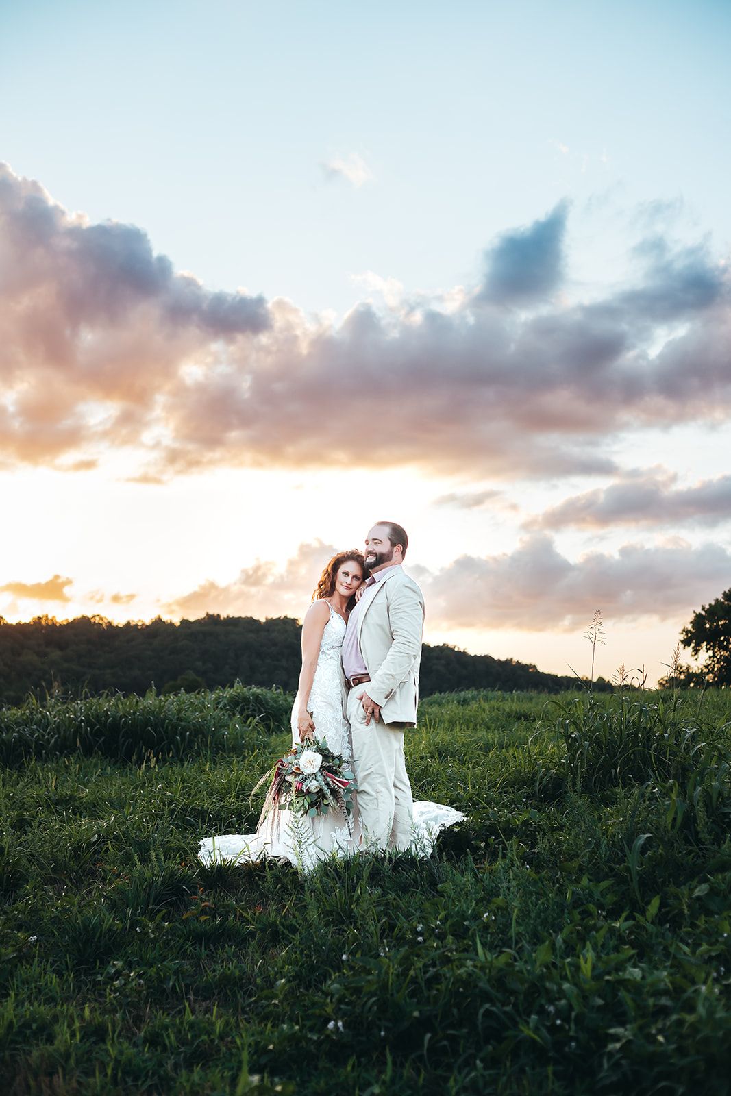 Newlyweds in white attire pose together in field during golden sunset.