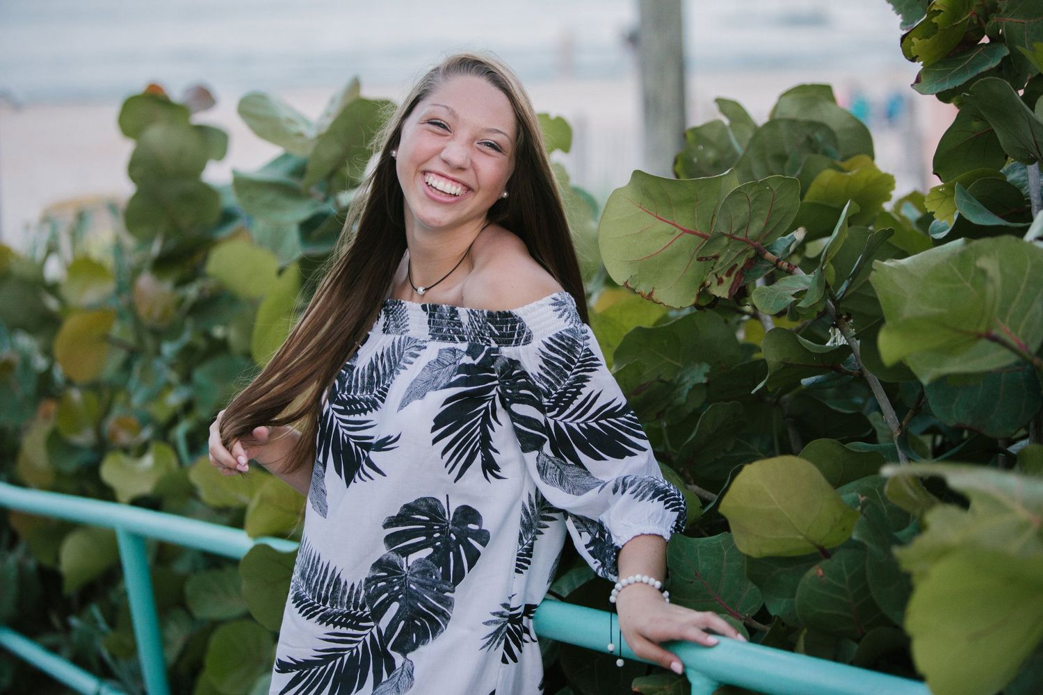 Woman in tropical print dress smiling by leafy green coastal foliage.