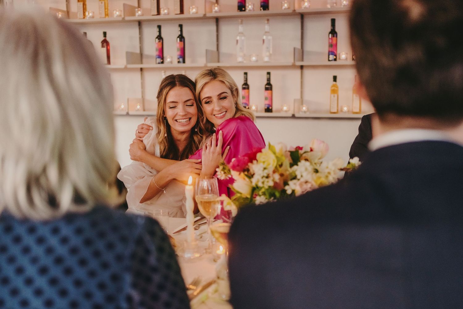 Wedding guests sharing laughter at reception dinner with bottle display wall in background.