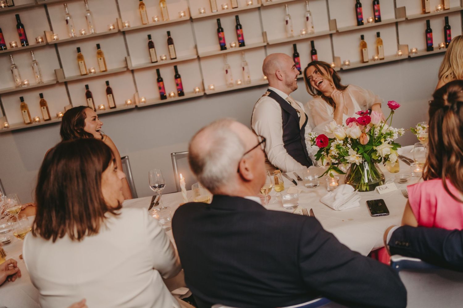 Wedding reception dinner with guests seated at tables against a wall of wine bottles.