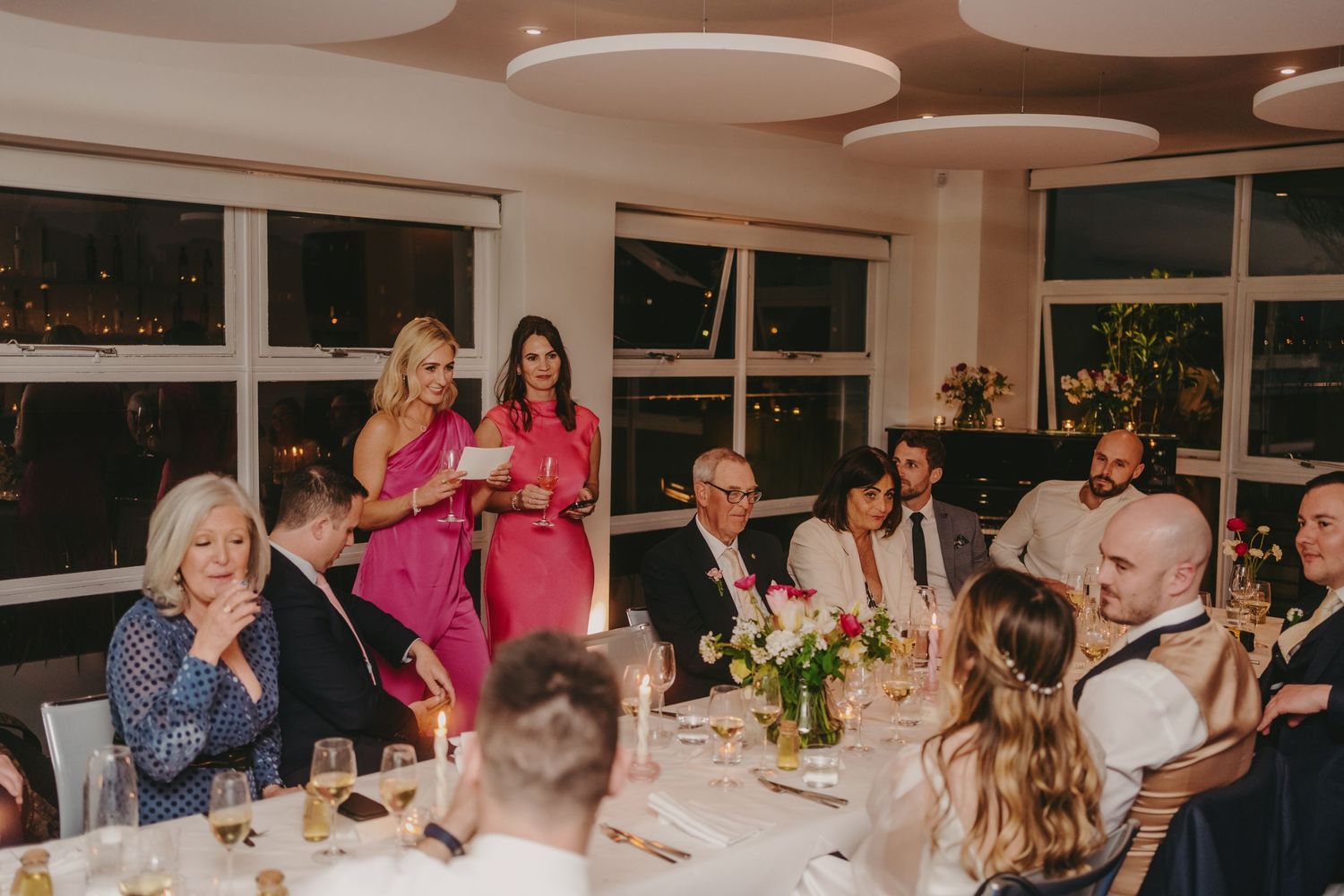 Wedding guests listening to reception speeches in an elegant dining room with circular ceiling lights.