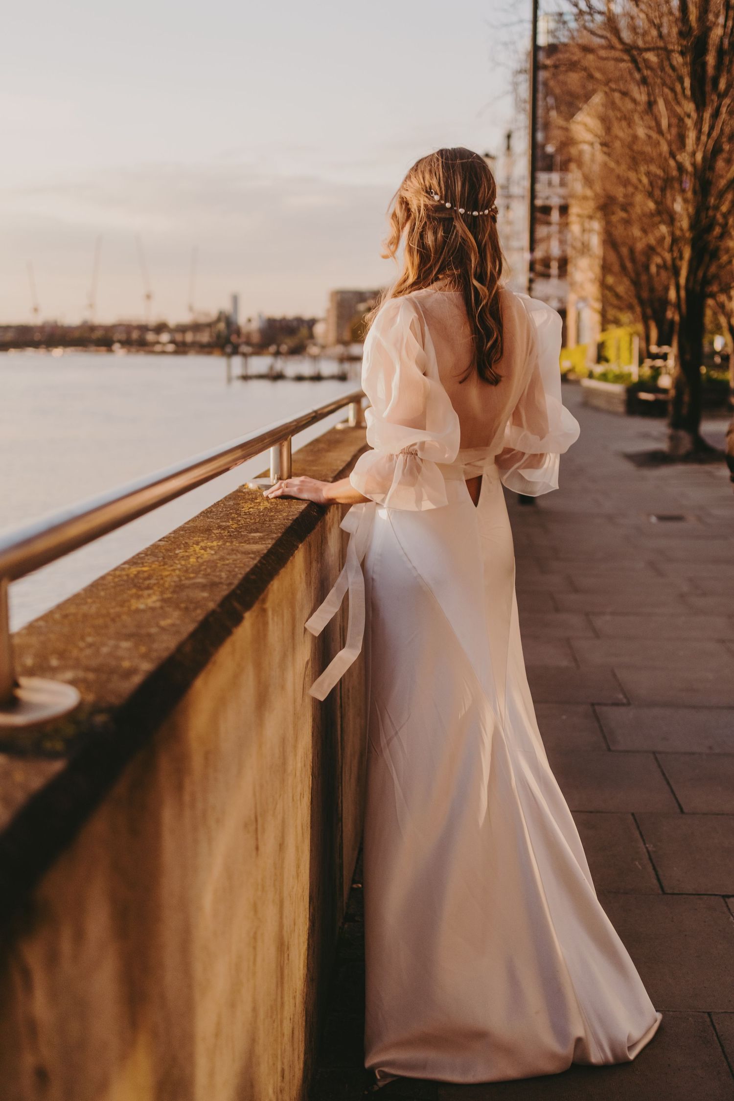 An elegant bride in a flowing white dress looks out over the waterfront at sunset.