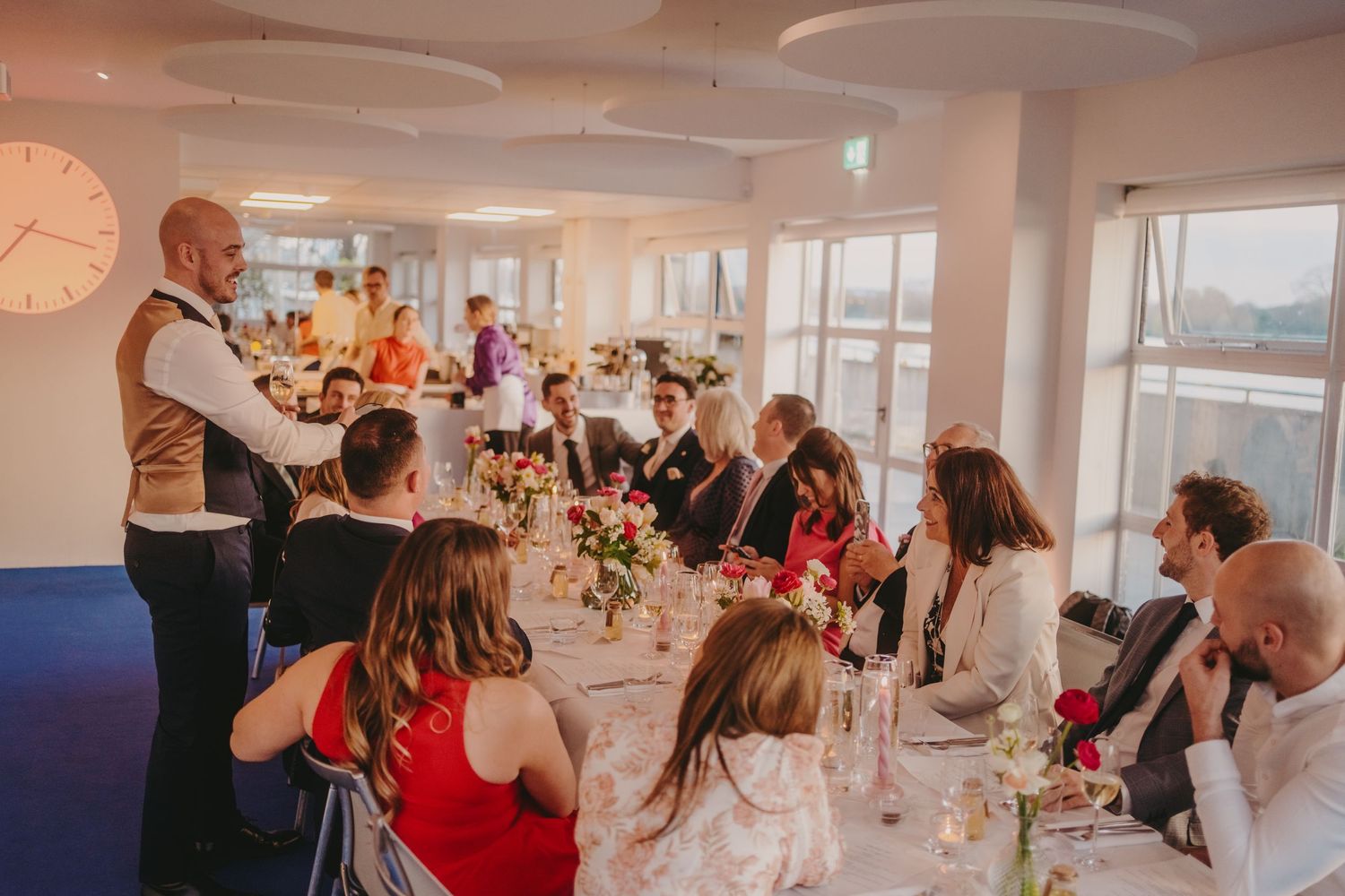 Guests gathered at an elegant wedding reception dinner with floral centerpieces and a waiter giving a speech.