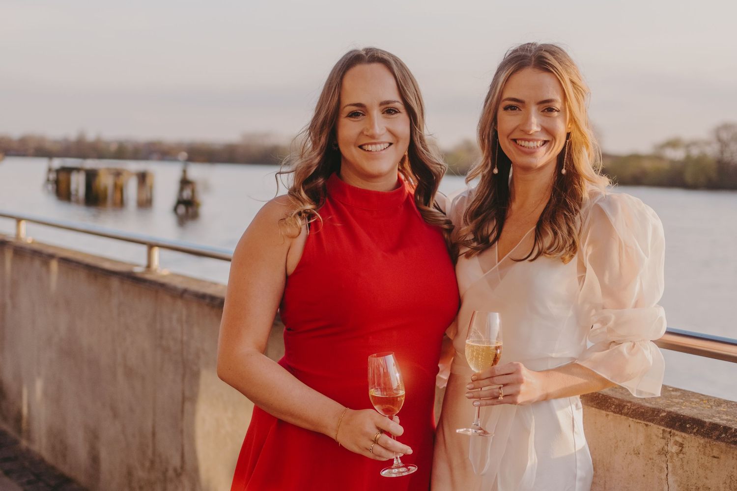 Two guests in red and white dresses pose with wine glasses at a waterfront event.