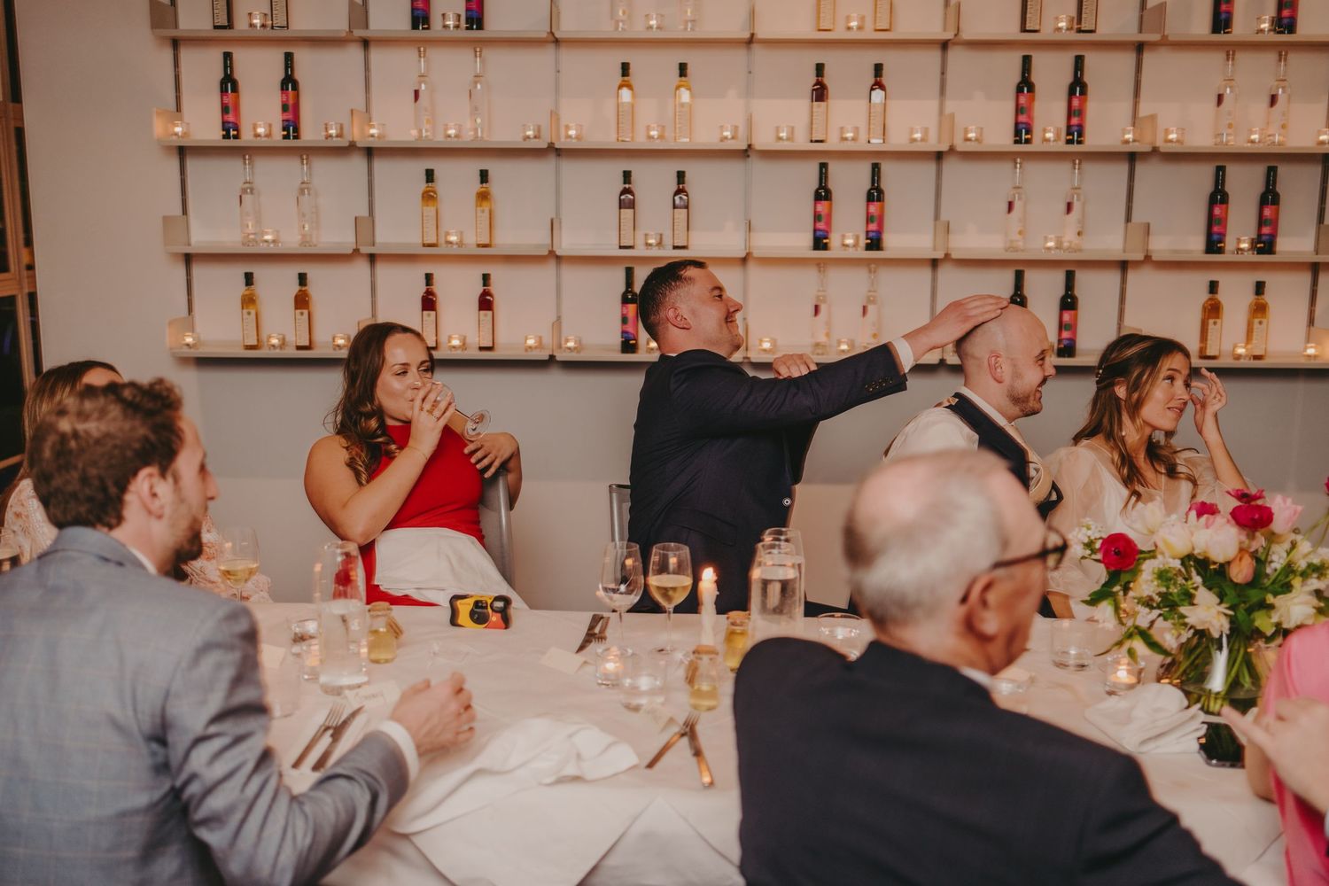 Animated dinner party scene with guests gesturing and laughing at a table in front of bottle-lined walls.