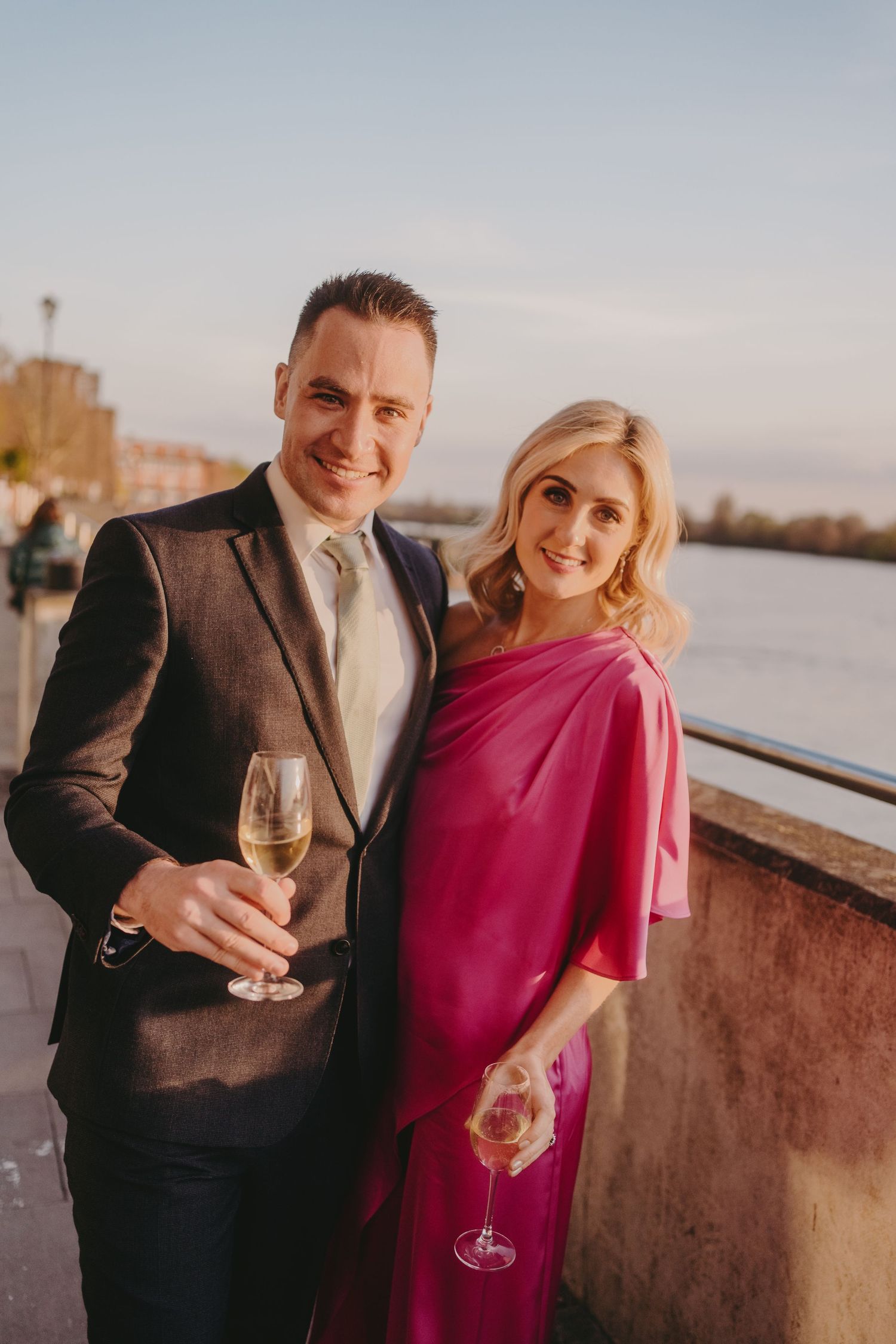 A couple in formal attire poses with champagne glasses along a riverside walkway.