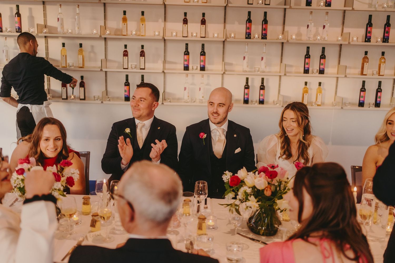 Formal dinner gathering with wine bottles displayed on white shelves in the background.