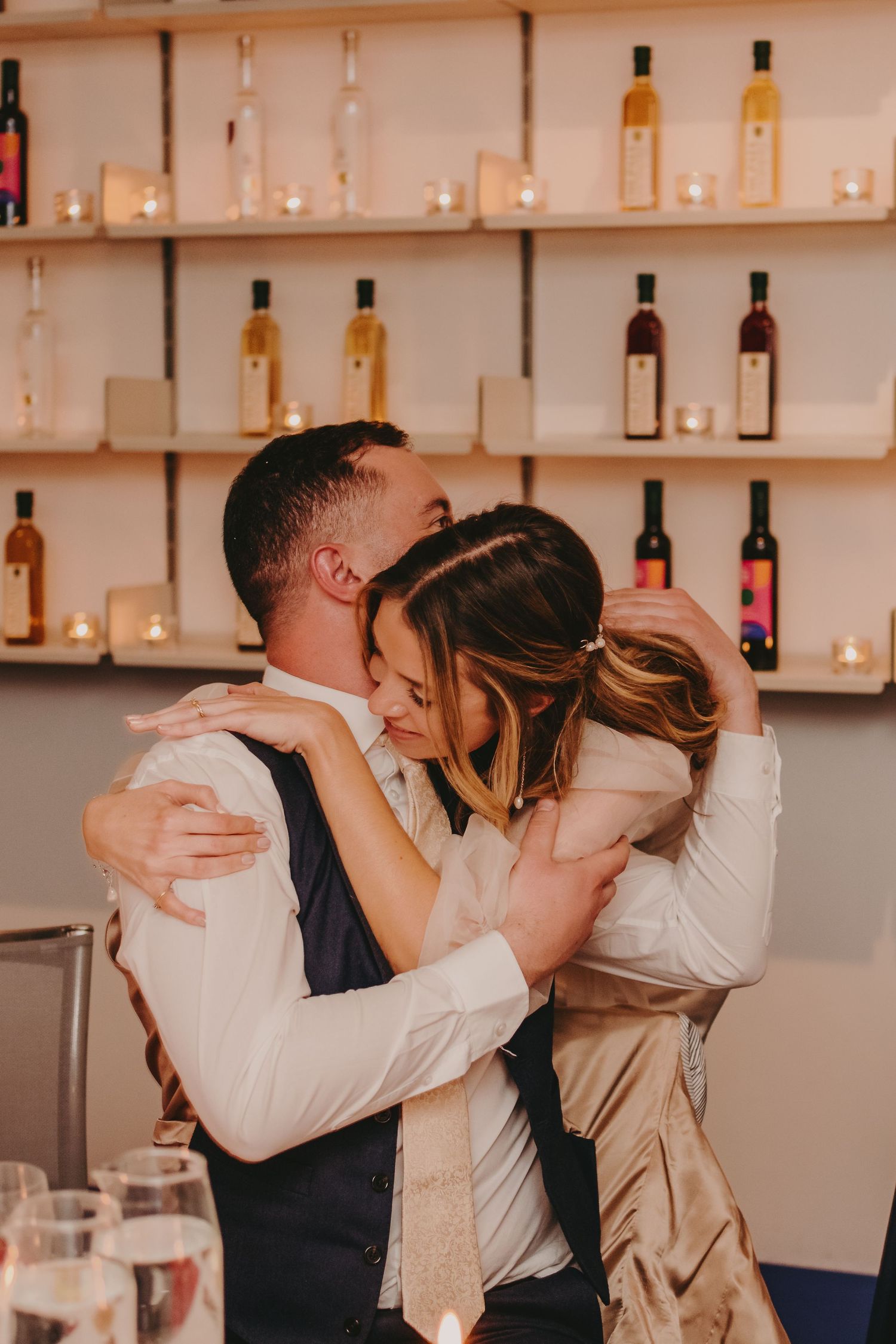 Romantic embrace between two people in formal attire against a backdrop of illuminated bottle shelves.