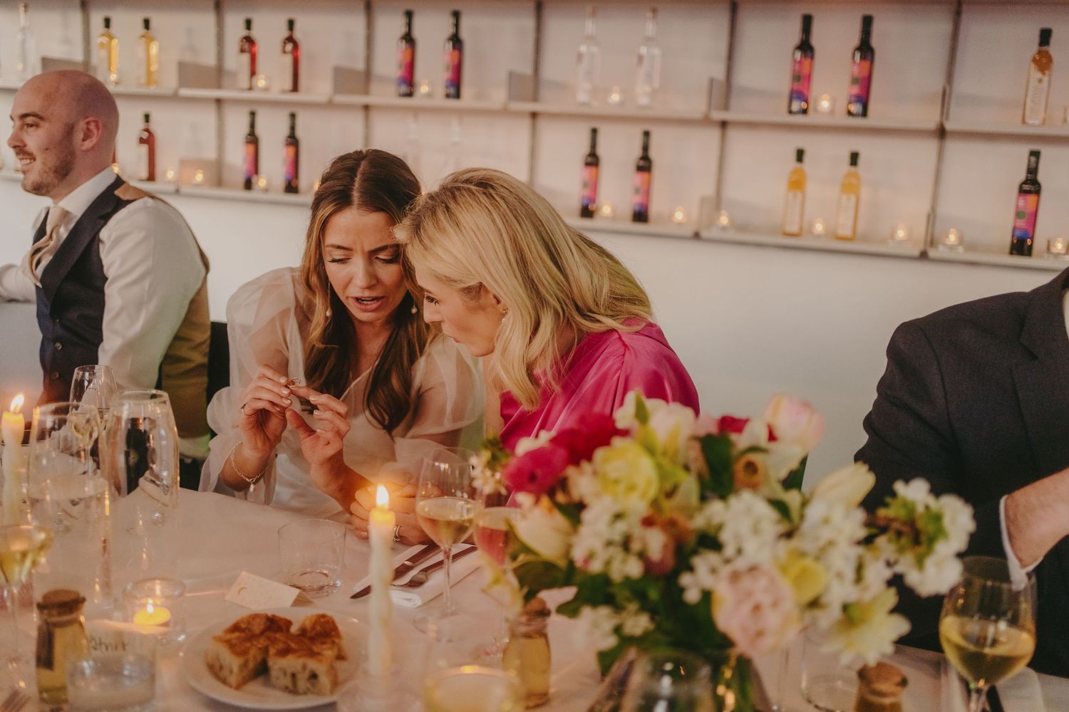 Guests enjoying wedding reception dinner with wine bottles displayed on shelves behind them.