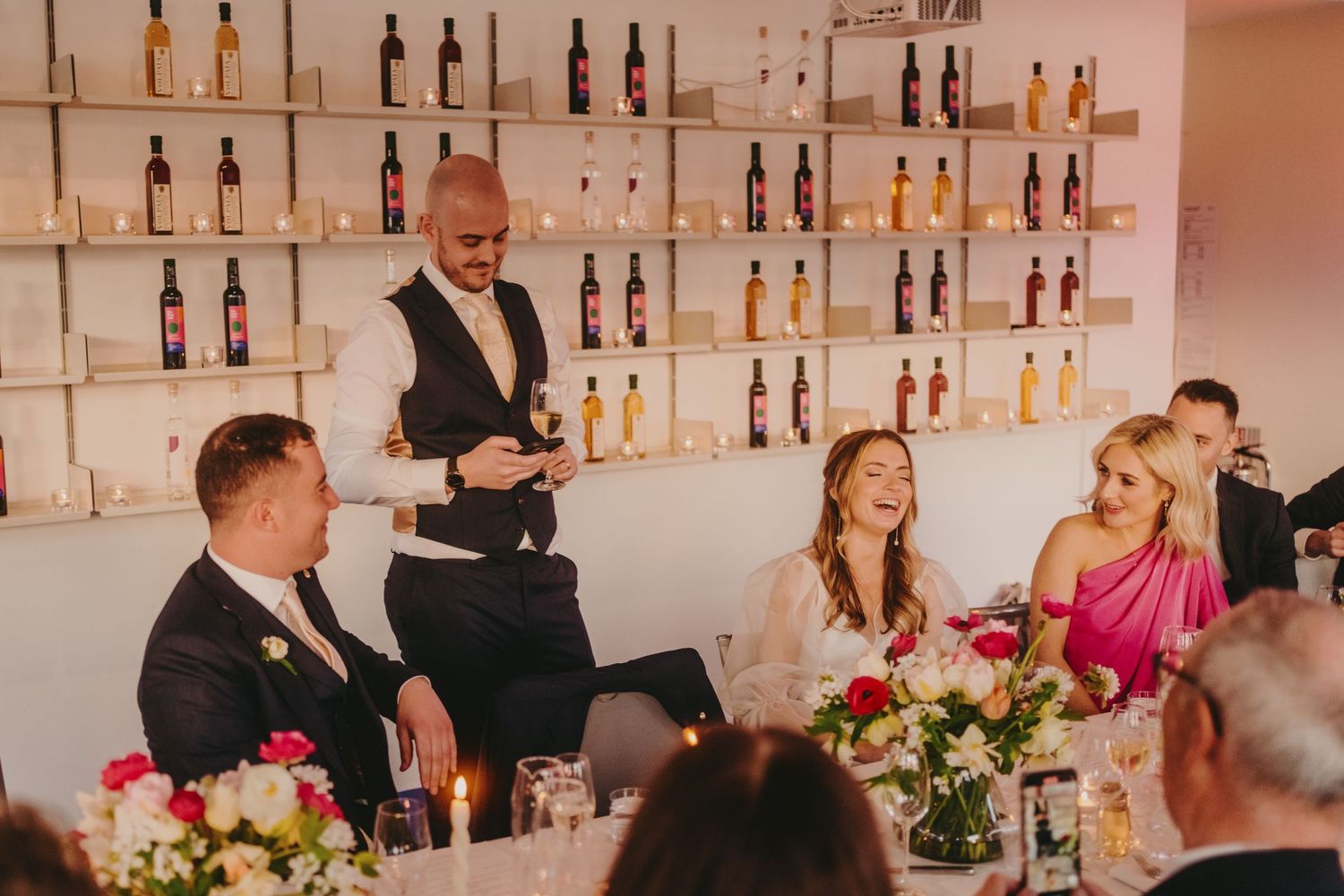 Wedding reception venue with shelves of bottles as guests dine at decorated tables.