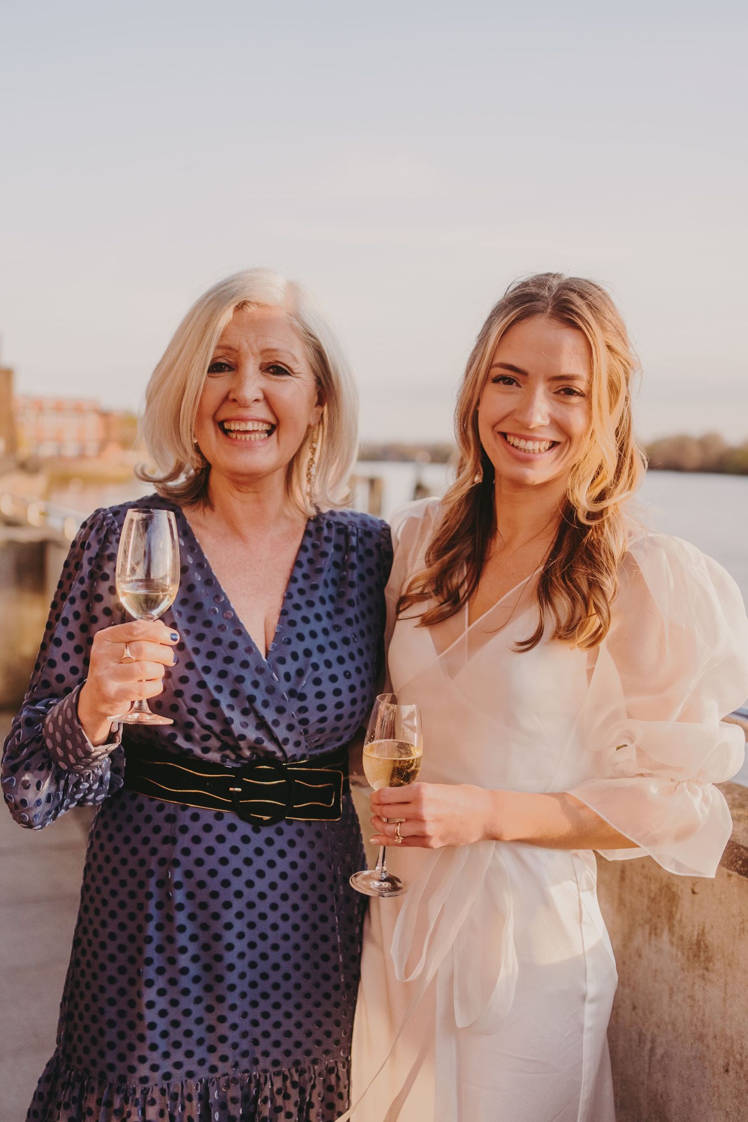 Two guests in polka dot and white dresses share a happy moment with champagne by the water.