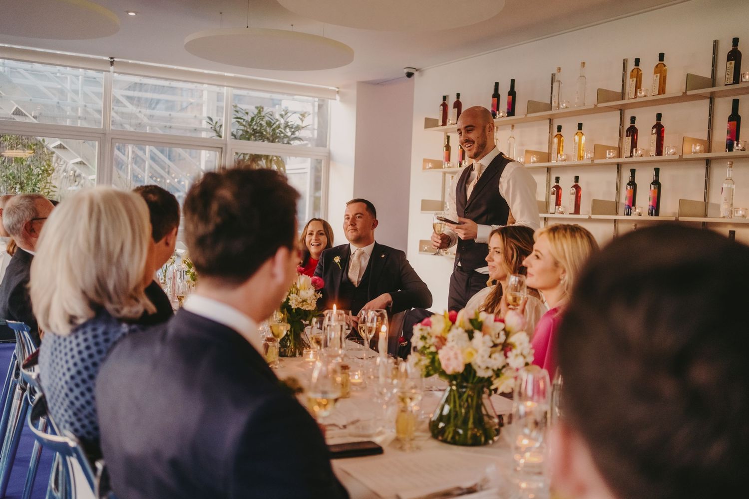 Modern restaurant interior with guests seated at long tables and decorative bottle displays.