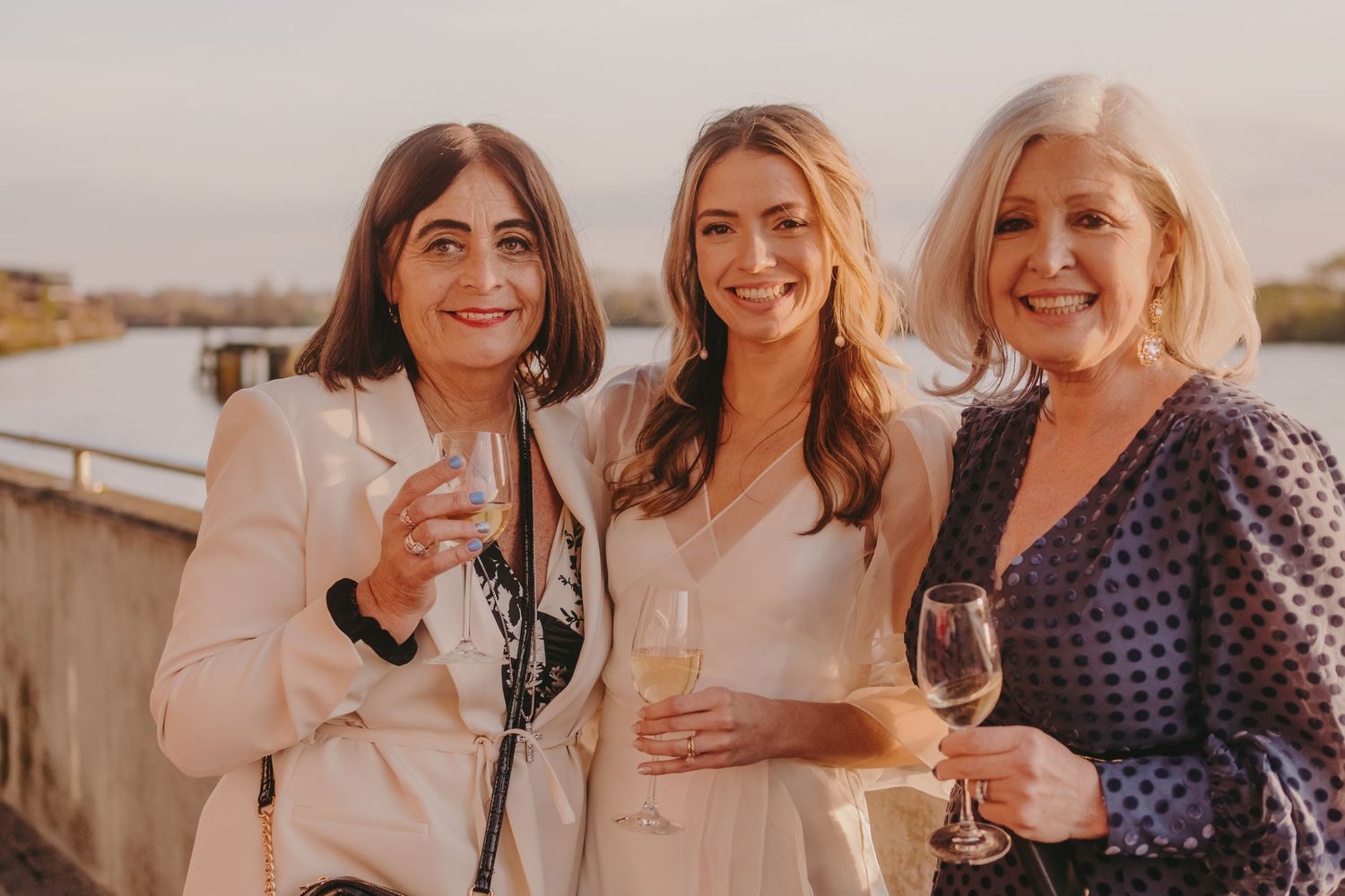 Three guests in formal attire pose with wine glasses at a waterfront celebration.