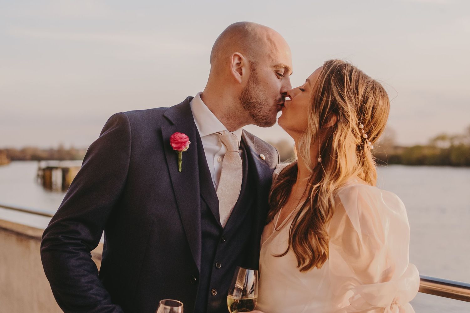 A romantic kiss between newlyweds against a waterfront backdrop at dusk.