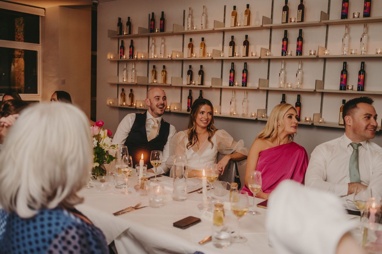 Guests enjoying a celebratory dinner at a restaurant with bottles displayed on shelves behind them.