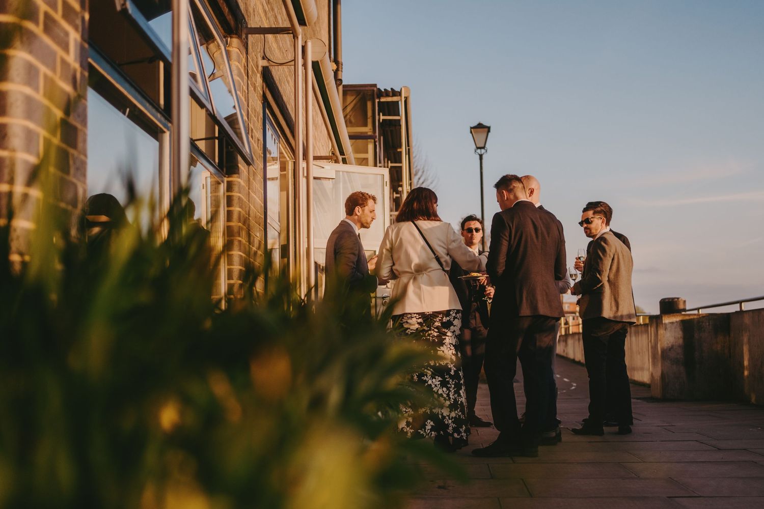 Guests gather on an outdoor terrace at sunset near a brick building with plants in the foreground.