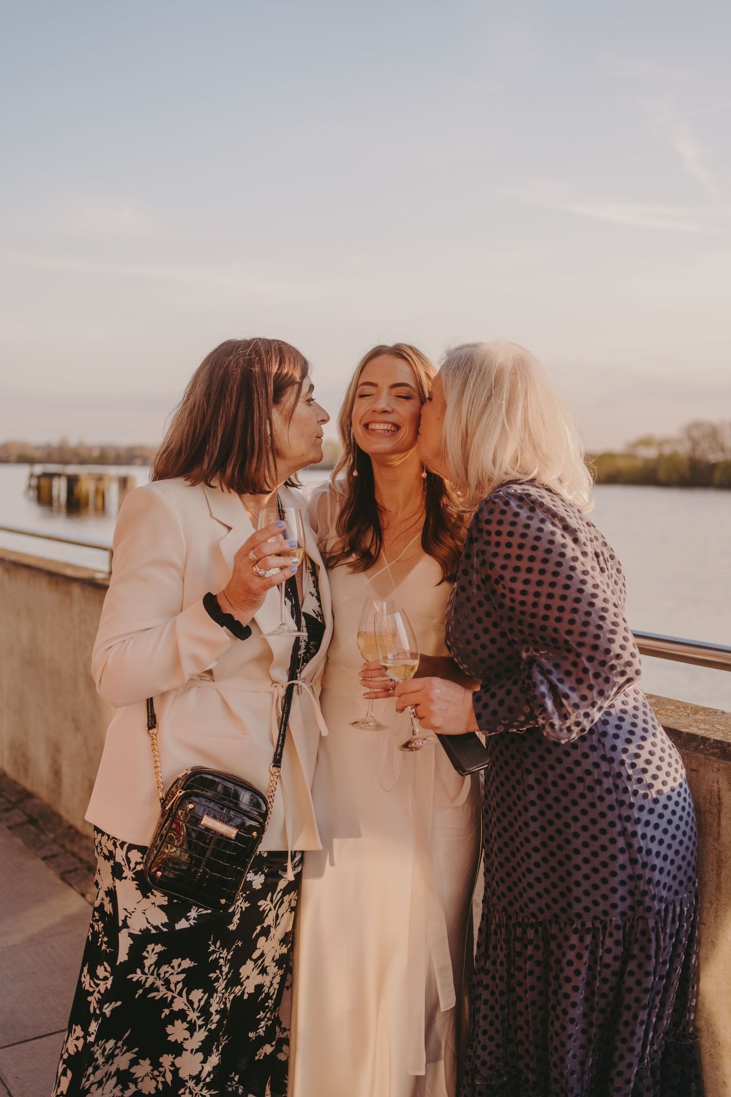Three friends share champagne and laughter at a waterfront celebration during golden hour.