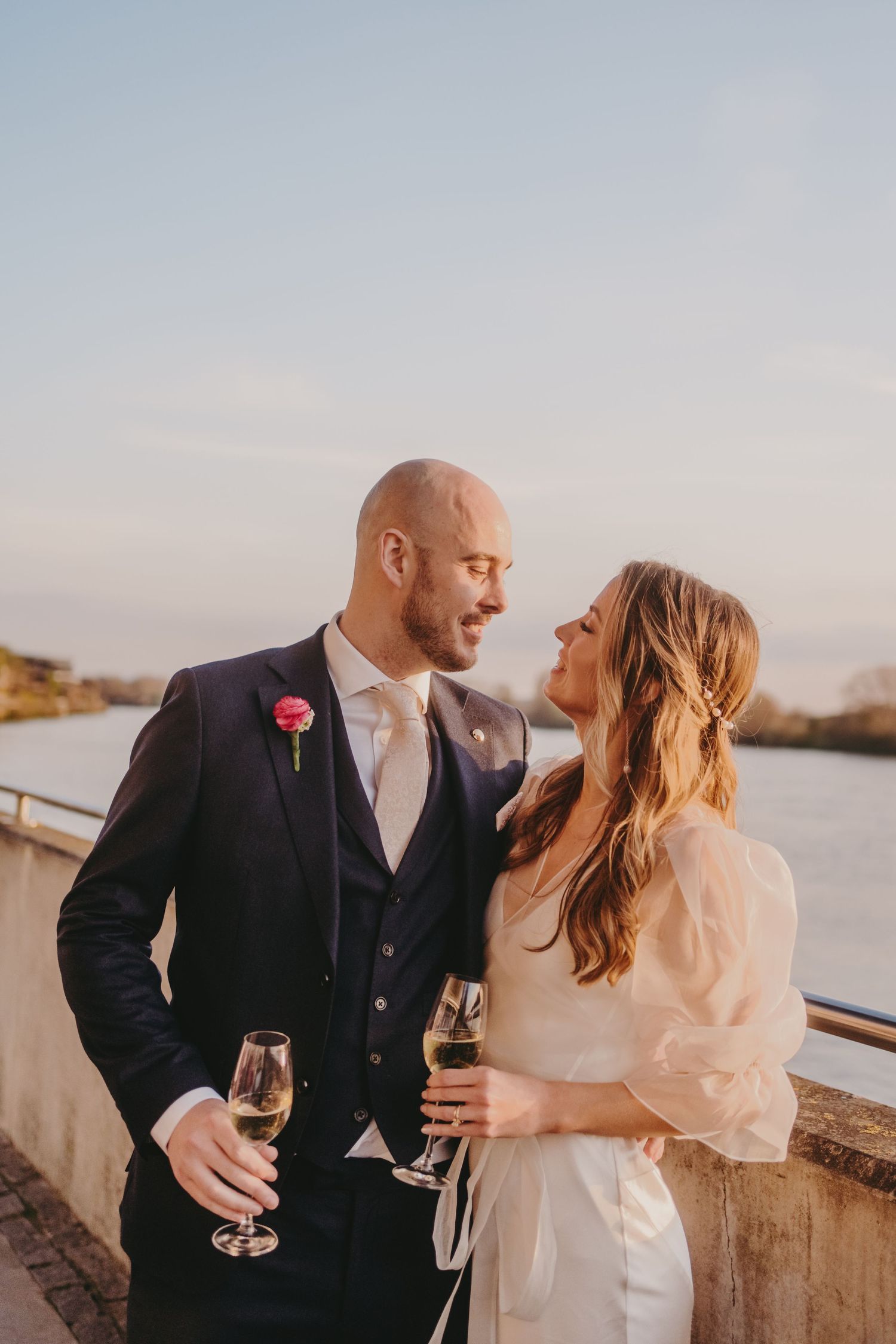 A wedding couple in formal attire shares an intimate moment while holding champagne glasses at sunset.