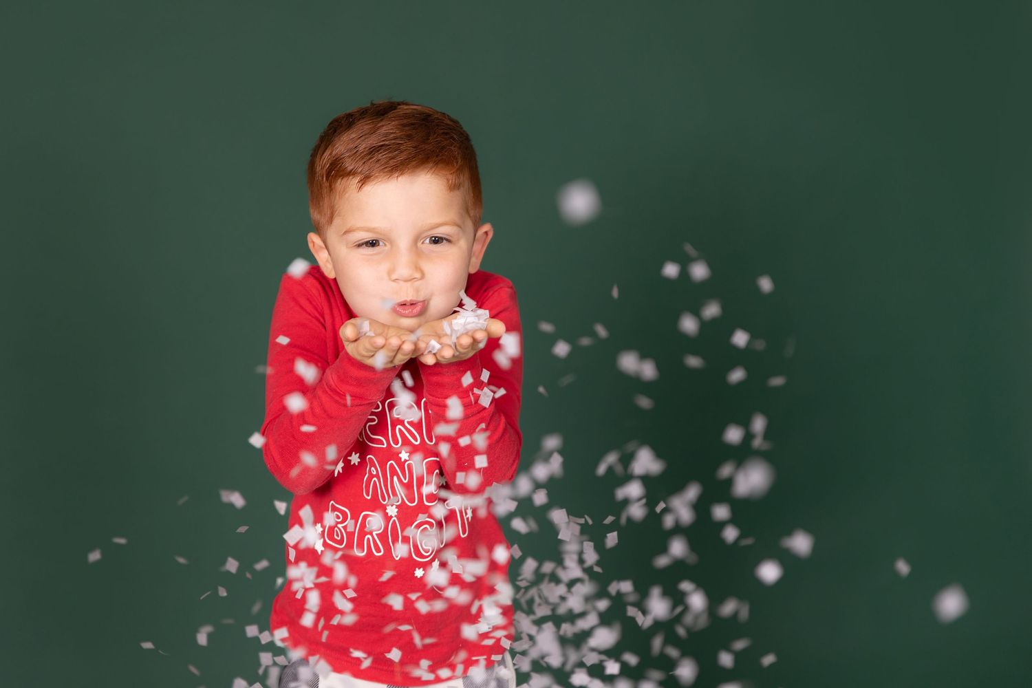 studio photo of child blowing fake snow out of hand wearing christmas pajamas on green backdrop studio mini clayton