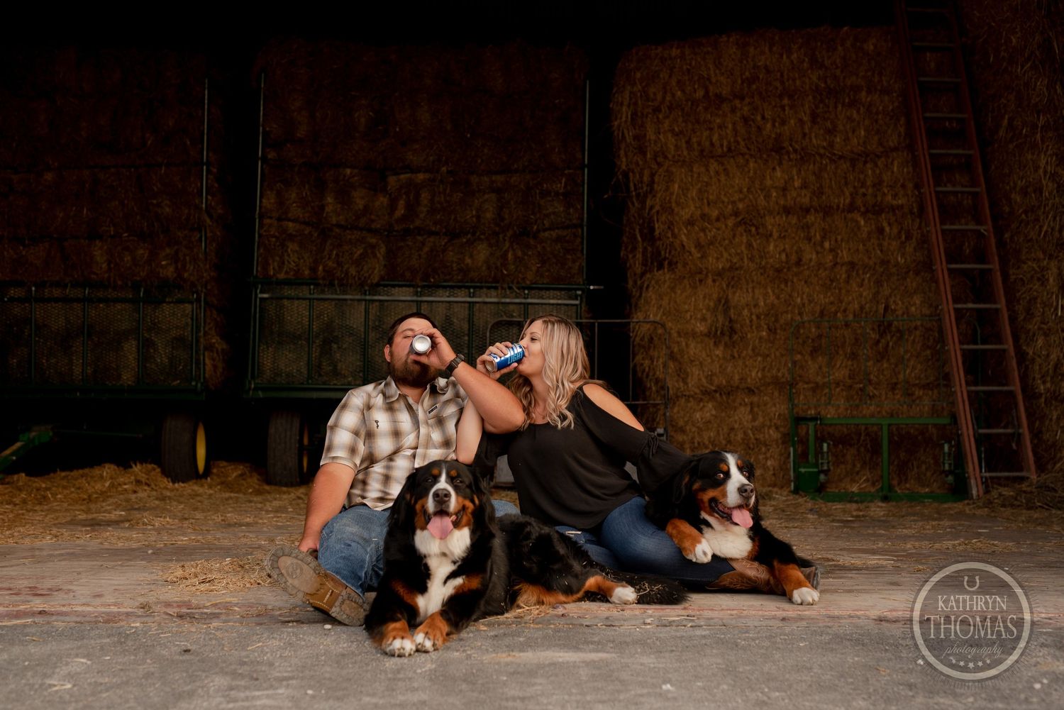 Couple relaxing with their Bernese Mountain Dogs in a rustic barn setting.