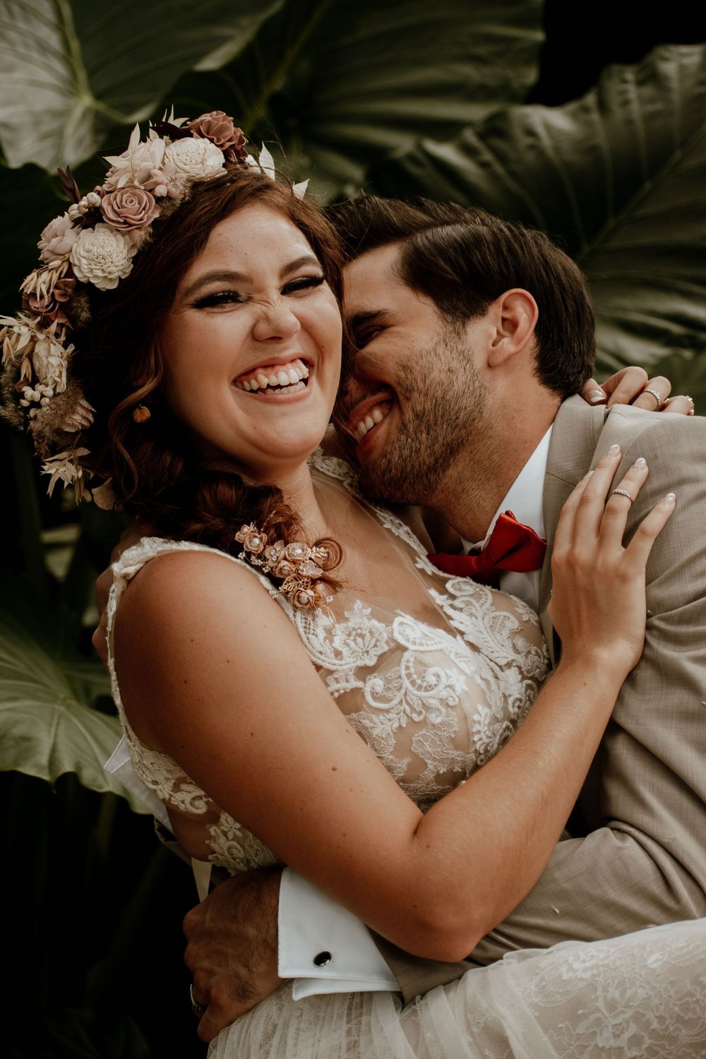 Joyful wedding couple sharing a tender moment with floral crown and beige suit.