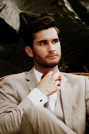 Groom in beige suit and red tie poses thoughtfully against dark background.