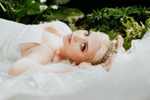Bride lying on white fabric surrounded by greenery wearing a delicate crown and elegant dress.