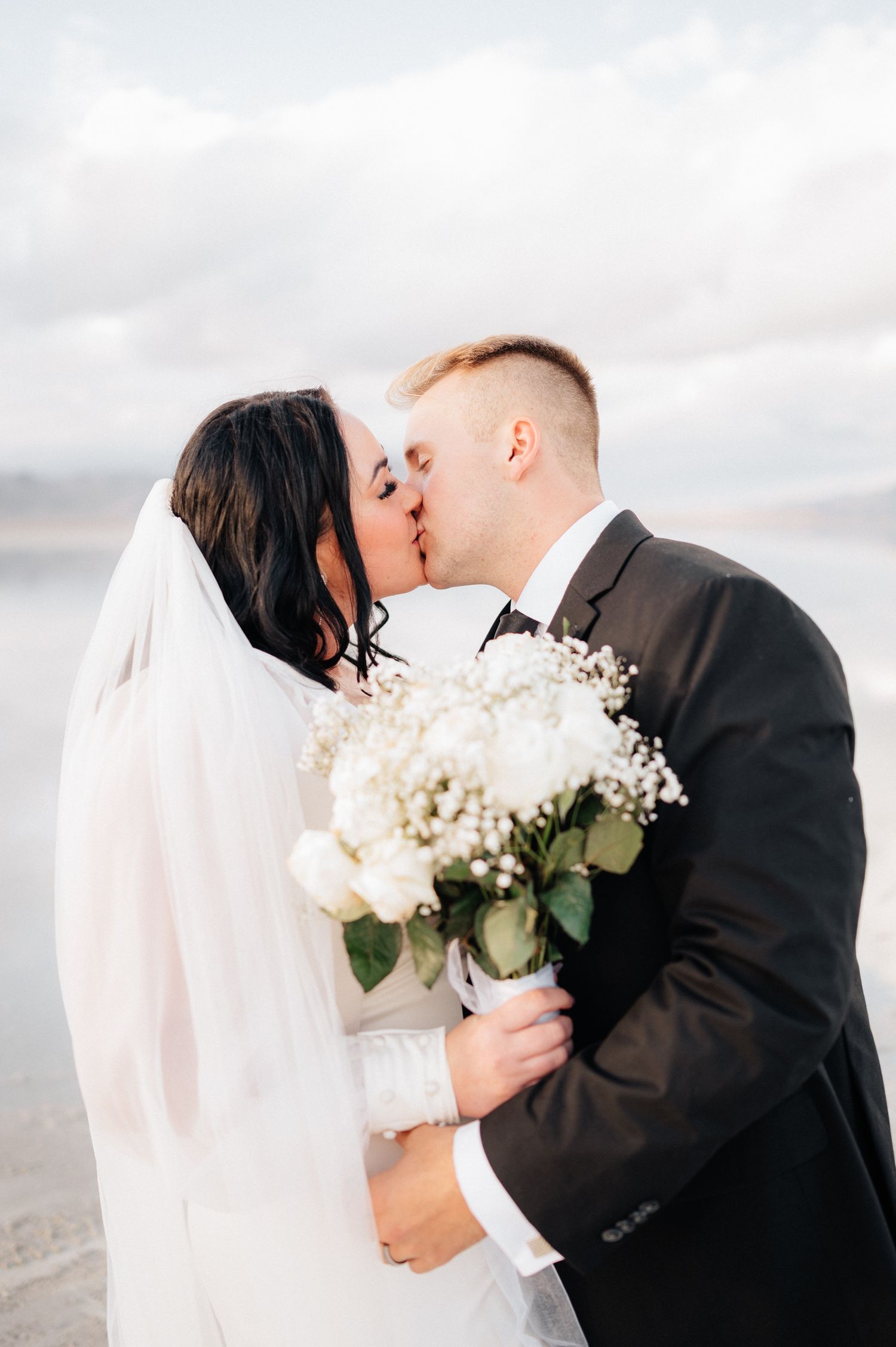 Romantic wedding couple shares kiss while holding white floral bouquet.