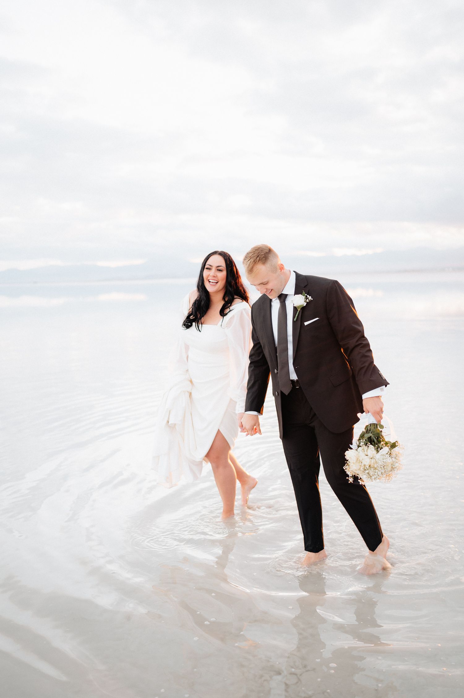 Newlyweds wade through shallow water at beach during overcast day.