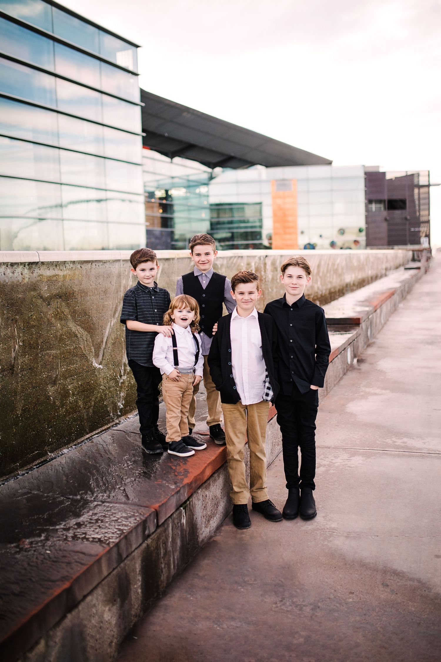 Group of five boys posing outdoors near modern glass building on urban walkway.