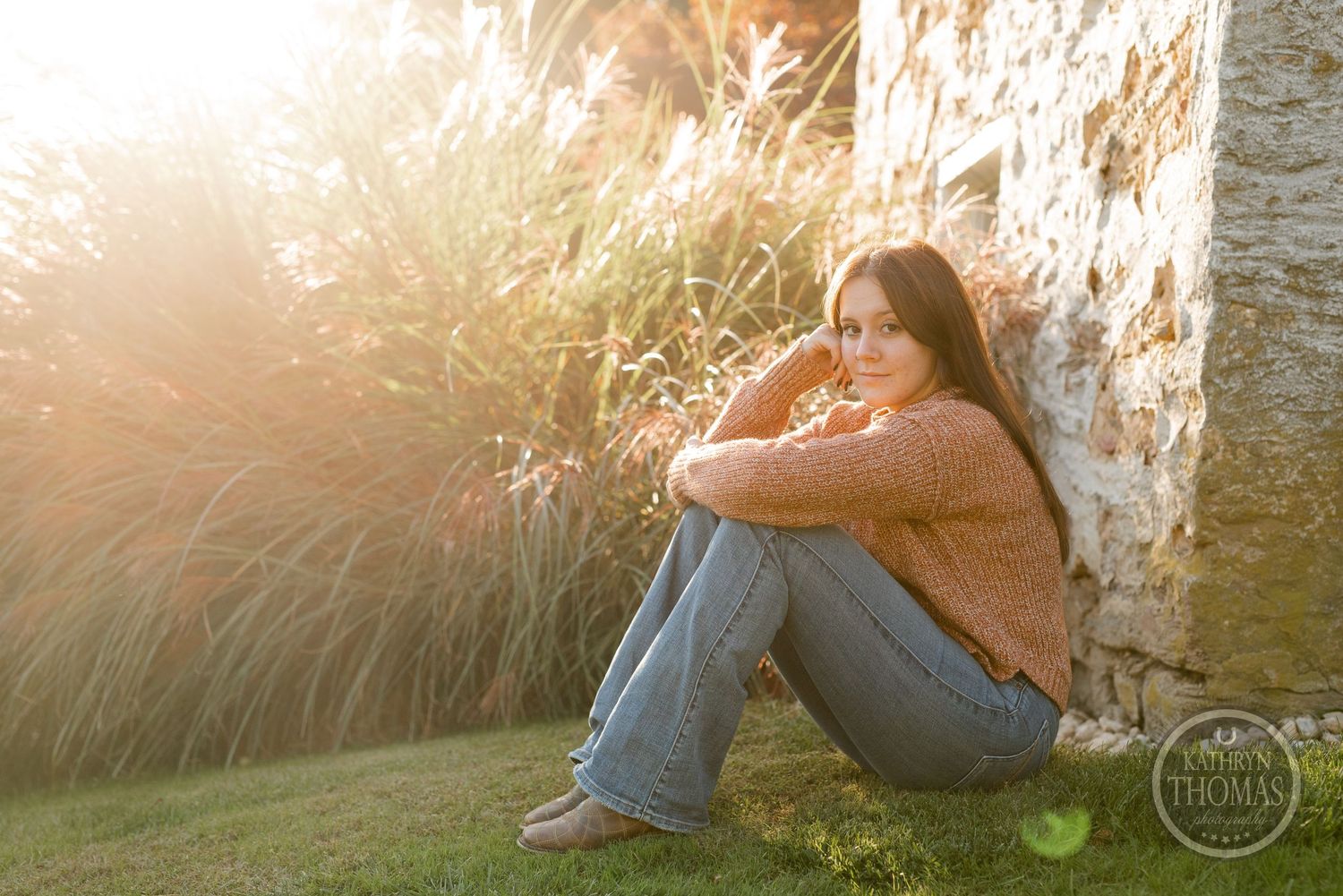 Someone in cozy sweater sits against stone wall during golden hour with tall grass nearby.