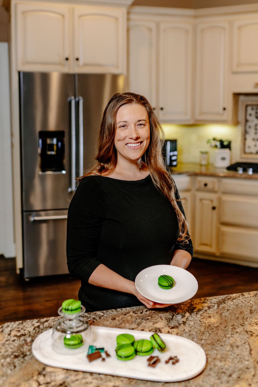 A baker preparing green macarons in a modern kitchen with cream cabinets.