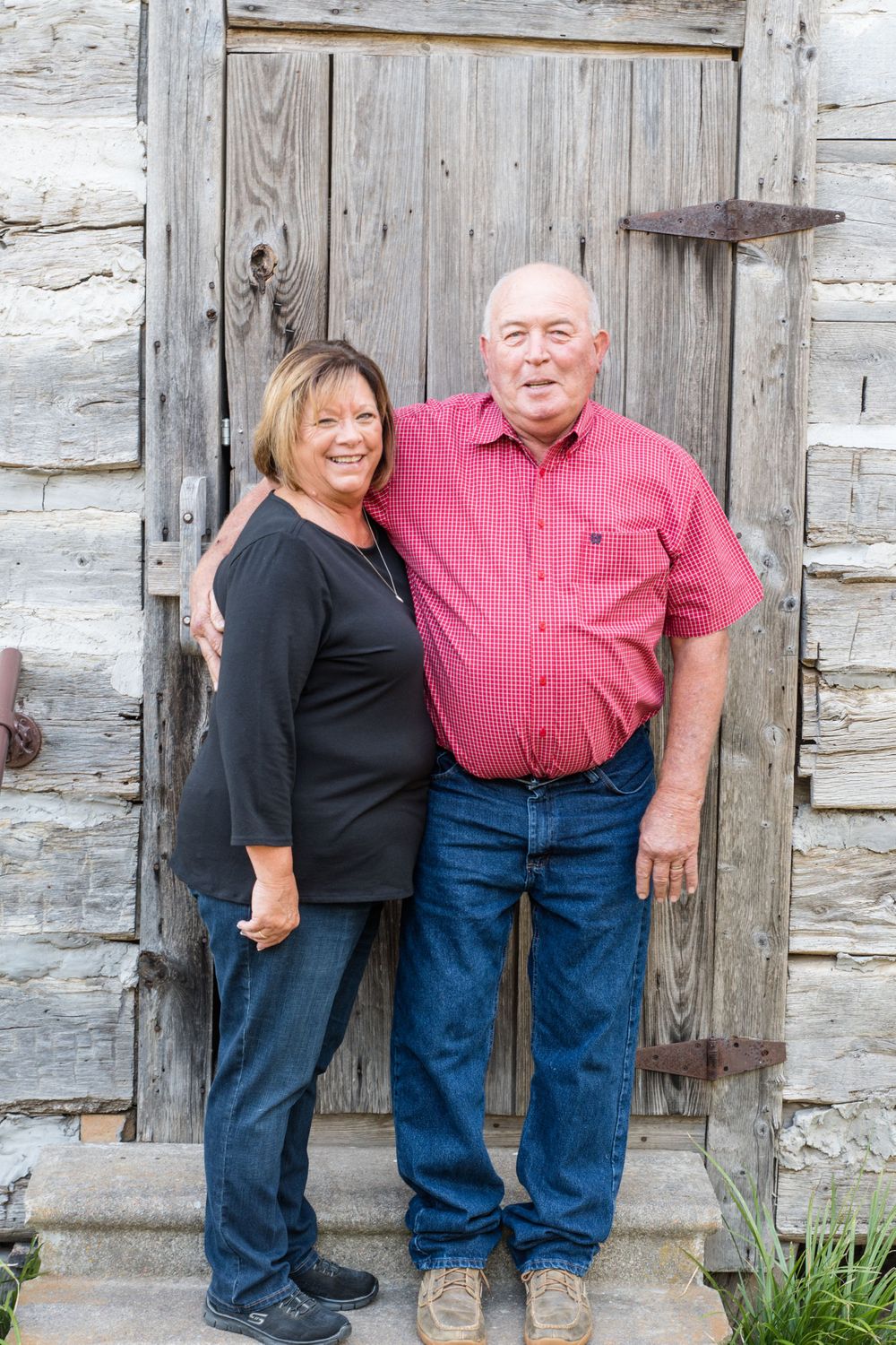 Couple poses in front of weathered wooden door on brick steps.