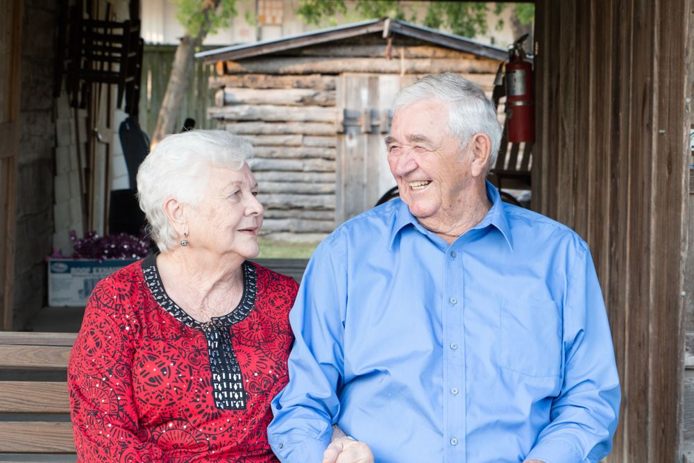 Elderly couple in red and blue sharing a loving moment on rustic porch.