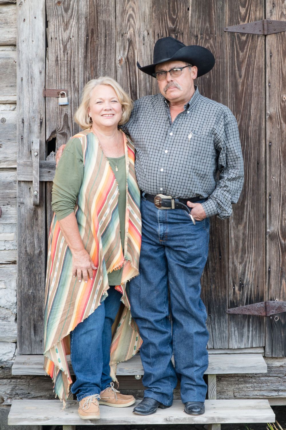 Couple in western wear standing on wooden porch step of rustic building.
