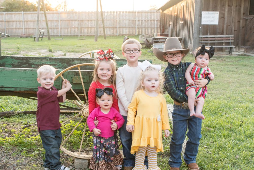 Group of young children in country attire pose by vintage wagon wheel in farm setting.