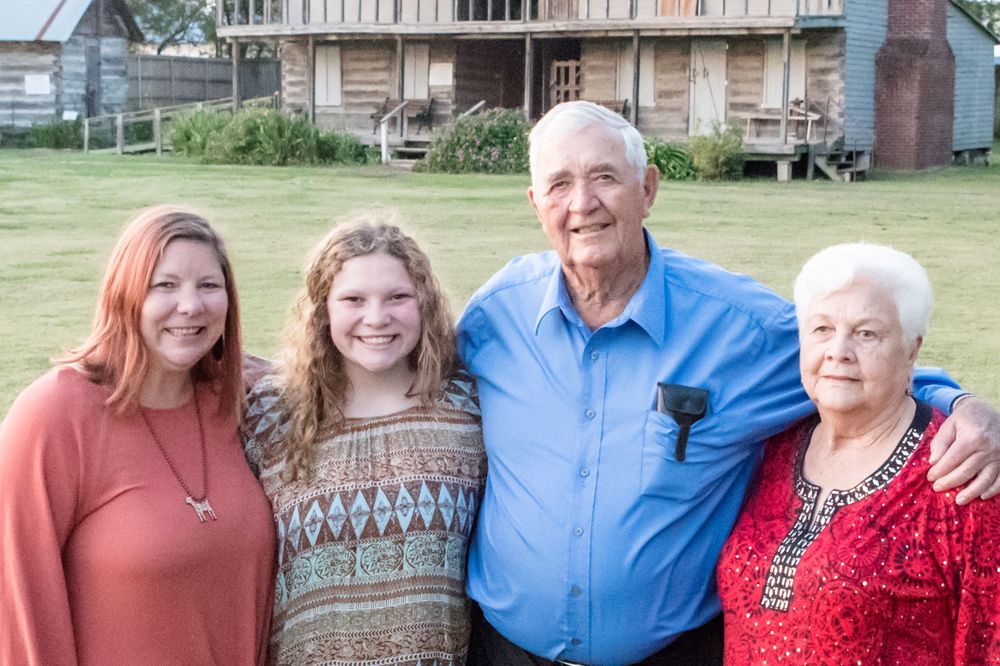 A casual outdoor family portrait taken on a sunny day with a rustic building in the background.