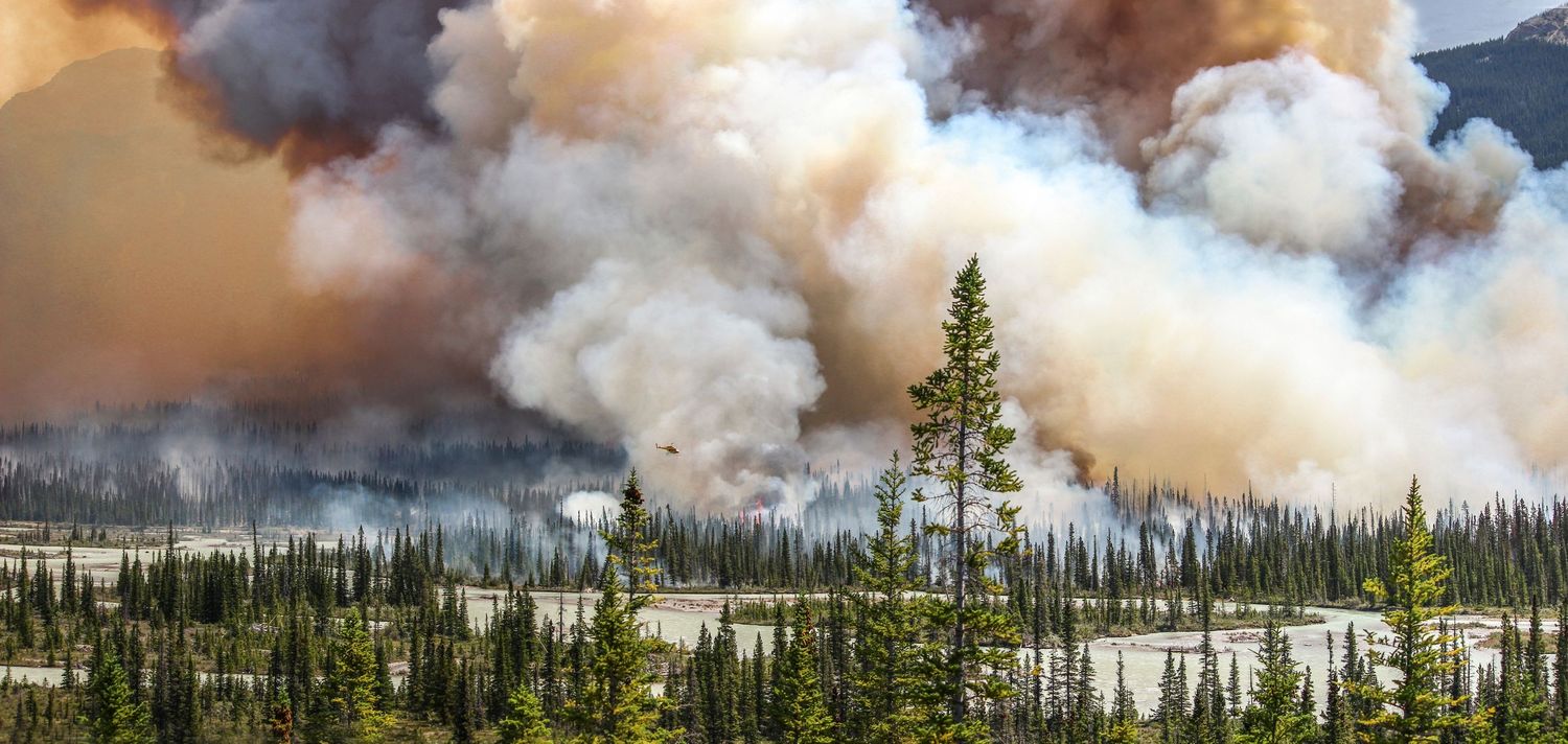 Massive wildfire sending huge smoke clouds over a boreal forest landscape with pine trees and river.