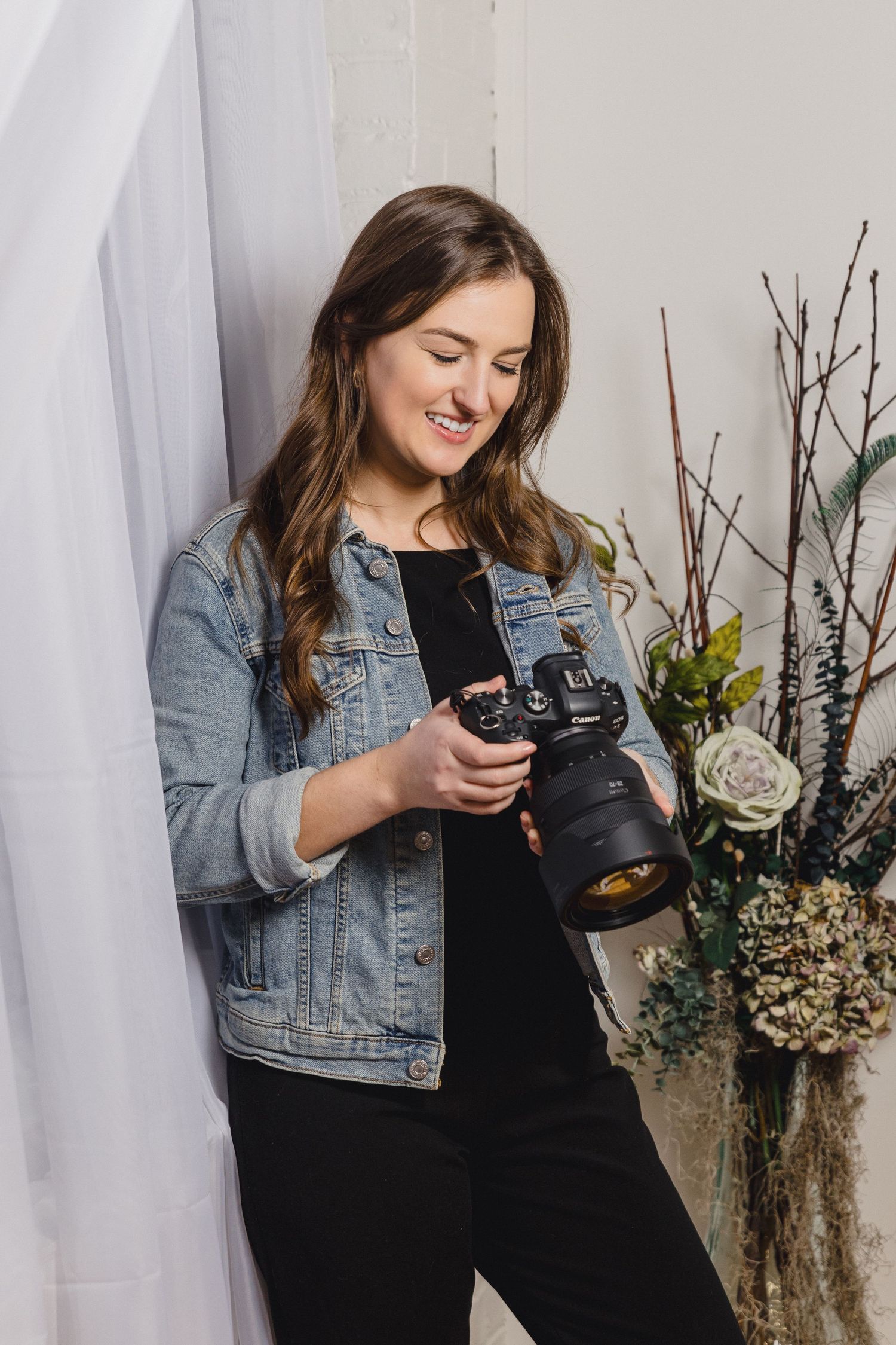 A photographer in denim jacket reviews images on their camera near floral arrangements.